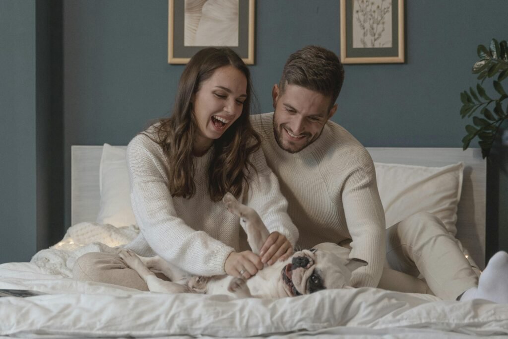 Couple laughing and petting their French bulldog on bed, capturing warmth and happiness.